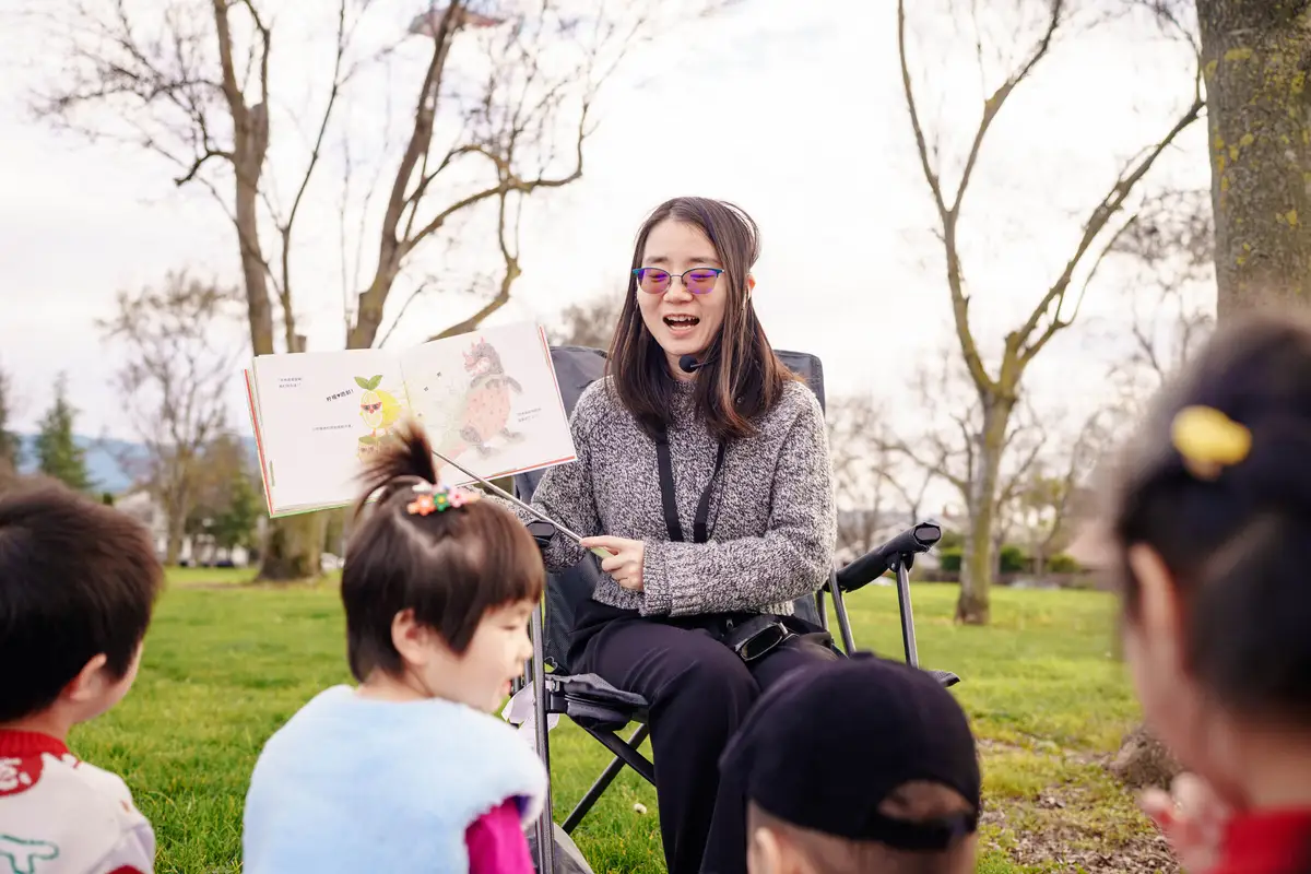 A storyteller reads an open Mandarin picture book aloud to children at an outdoor reading club gathering